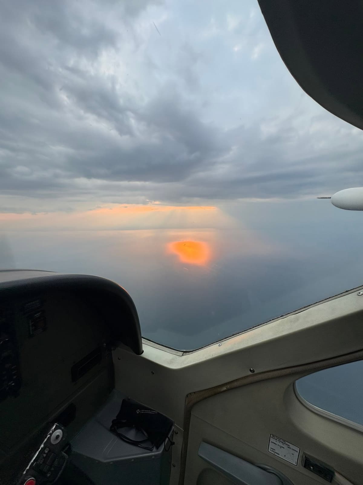 Sunset through the windscreen of a small aircraft, above a layer of cloud and calm water.
