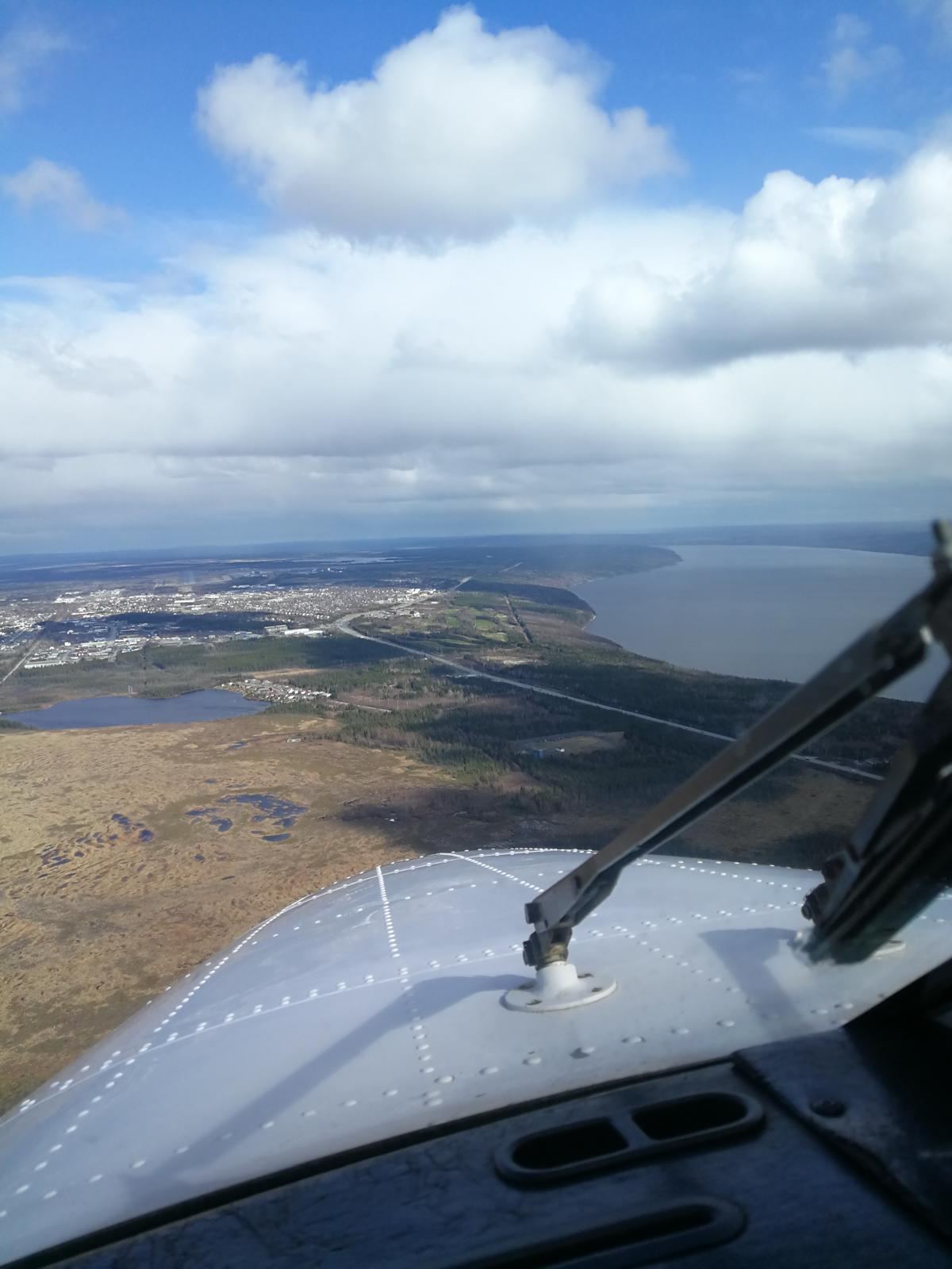 View over the nose of an aircraft toward a town beside a large lake, with forest below.
