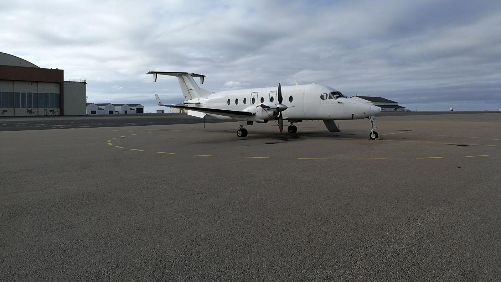 A white Beechcraft 1900D parked on a dark apron under an overcast sky.