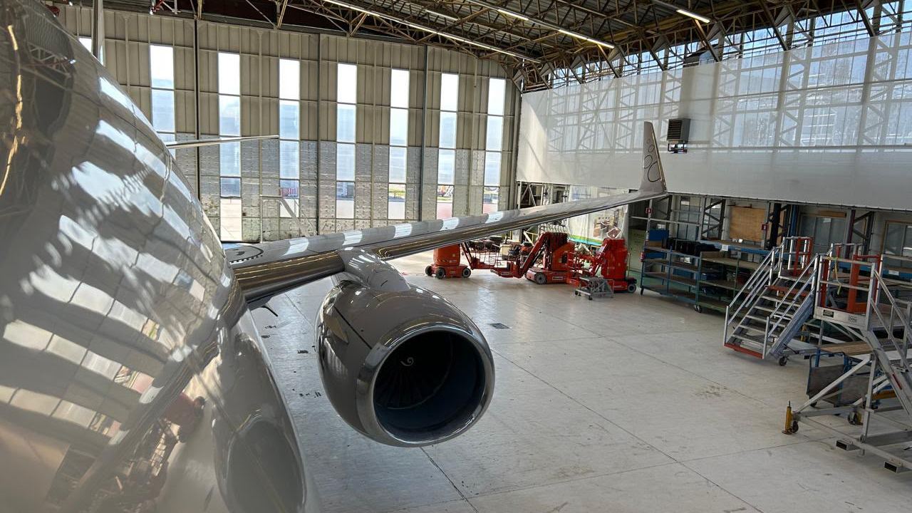 The wing, engine and winglet of a white Boeing jet in a hangar, seen from above.