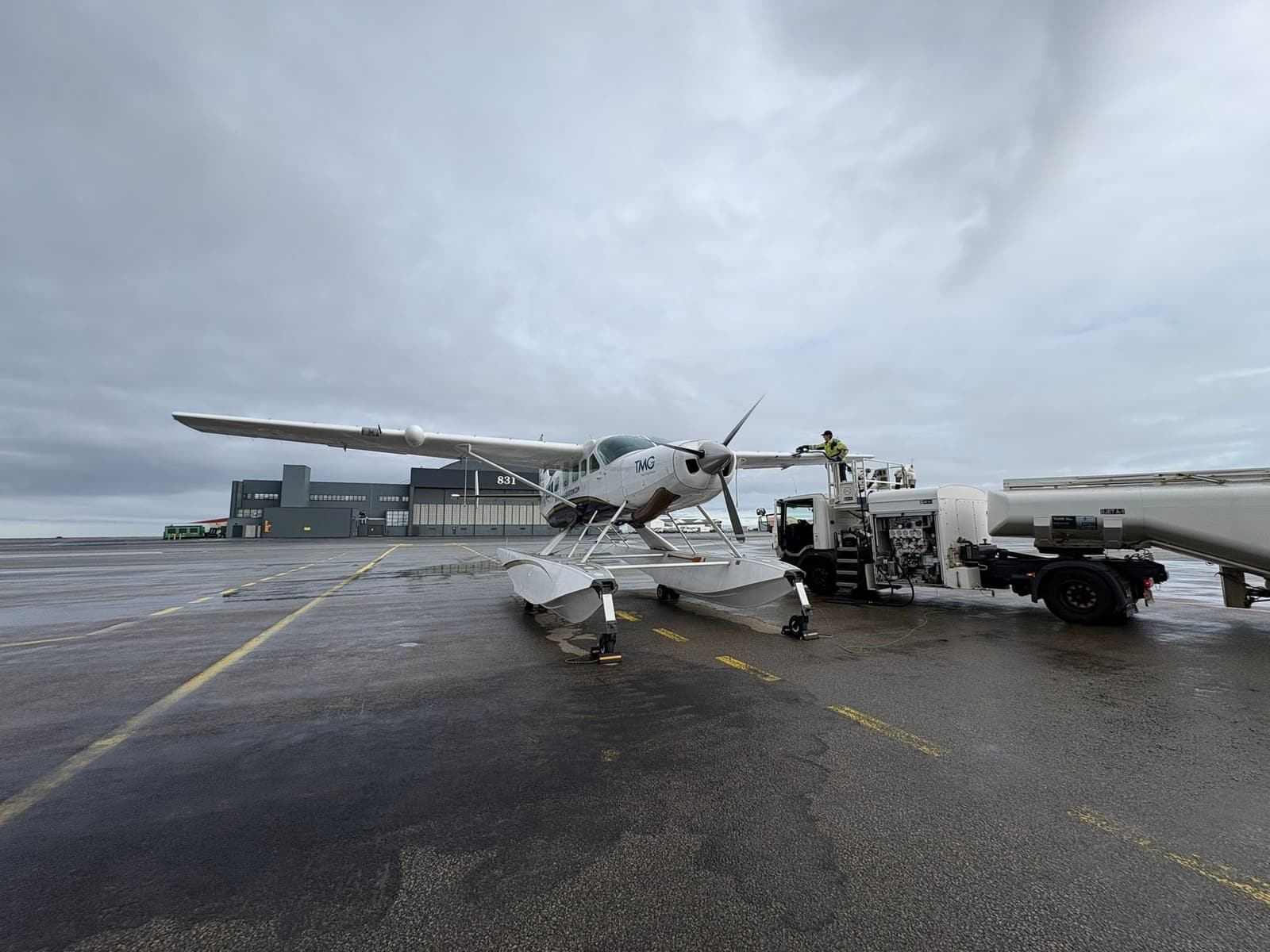 The amphibious Cessna Caravan being refuelled from a fuel truck on a wet ramp under an overcast sky.