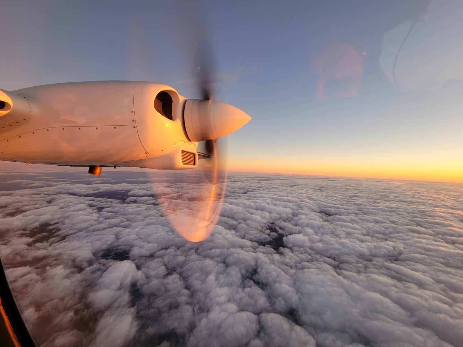 An engine and spinning propeller at sunset above the cloud, seen from the cabin.